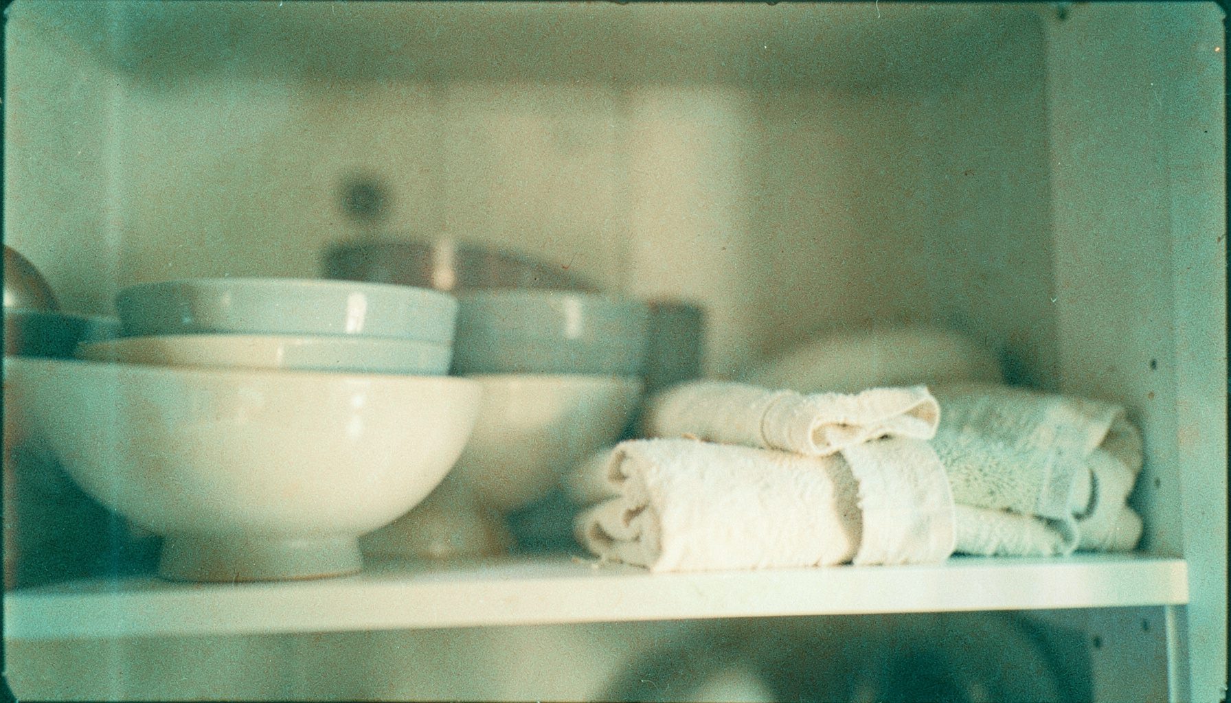 Analog film photograph of a softly lit kitchen shelf with stacked bowls and linen towels, blurred warm cream and sage tones, visible grain.
