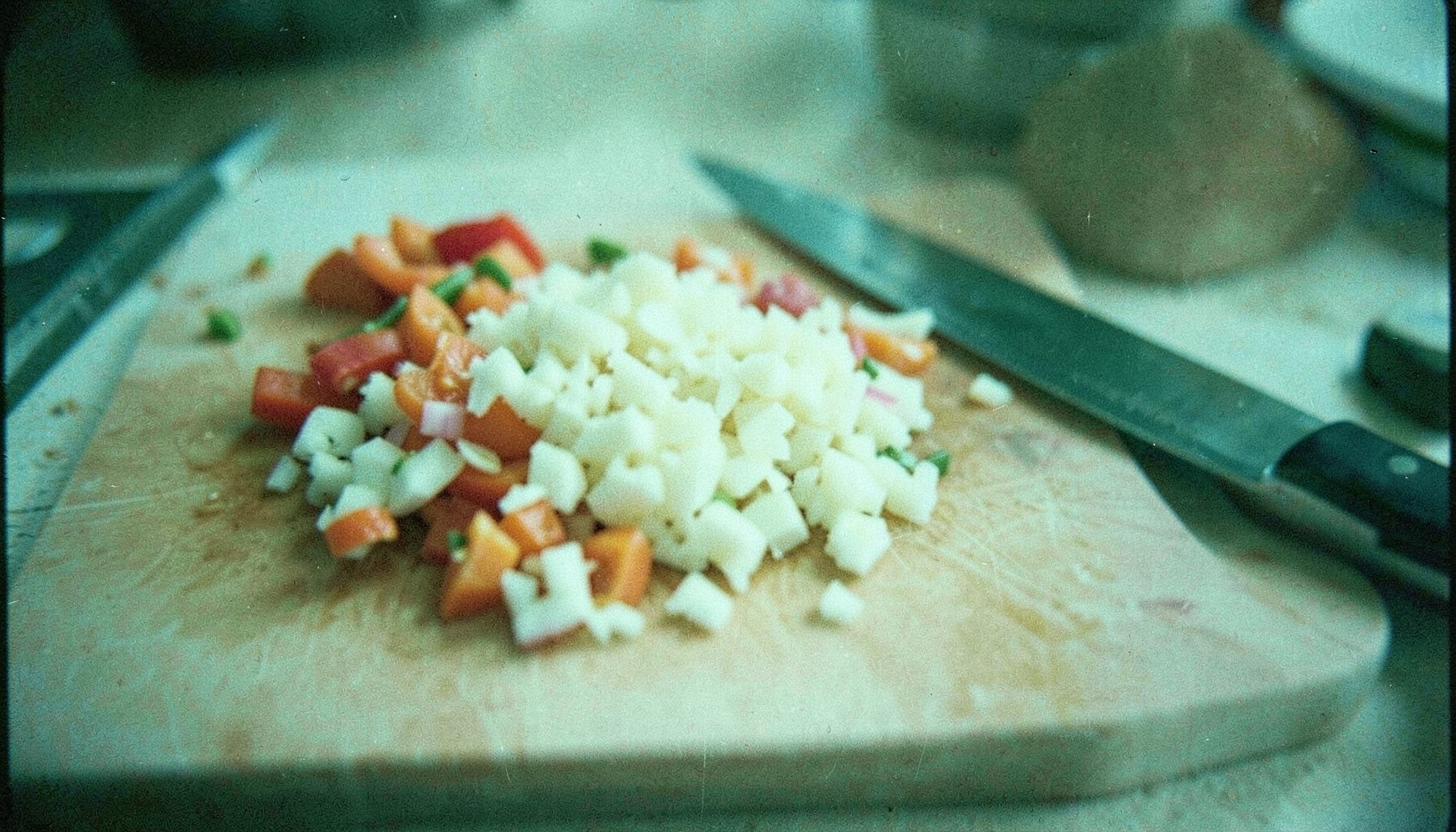 Analog film still of a cutting board with chopped vegetables and a chef's knife, natural daylight, visible film grain.