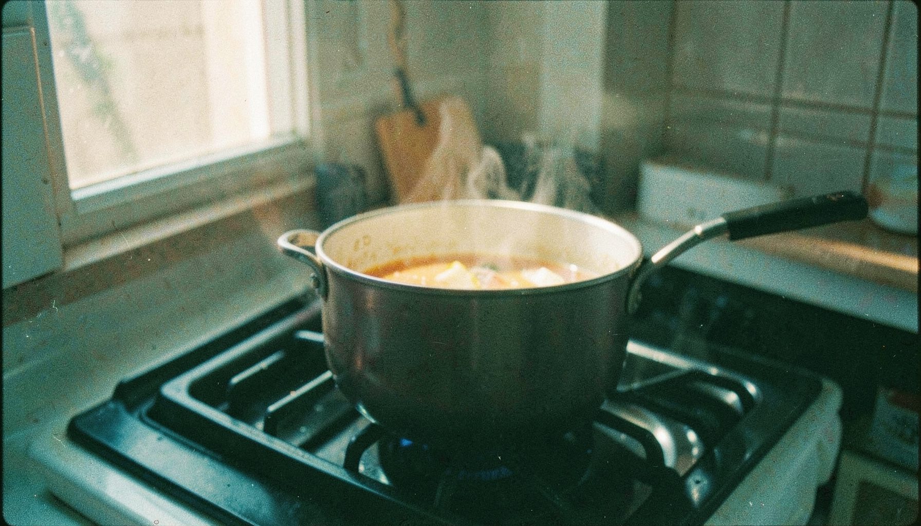 Analog film still of a stovetop with a simmering pot of soup, warm amber light from a kitchen window, visible grain.