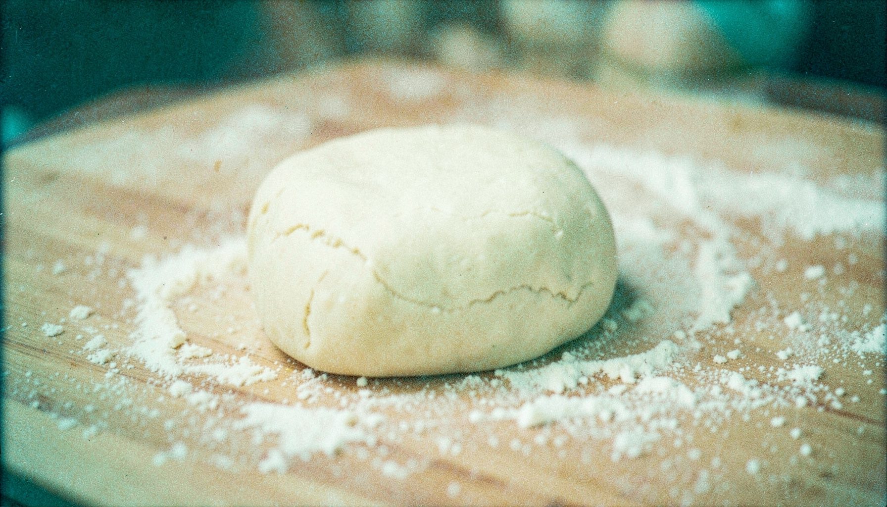 Analog film still of dough being rolled on a floured wooden surface, soft beige and cream tones, visible grain.